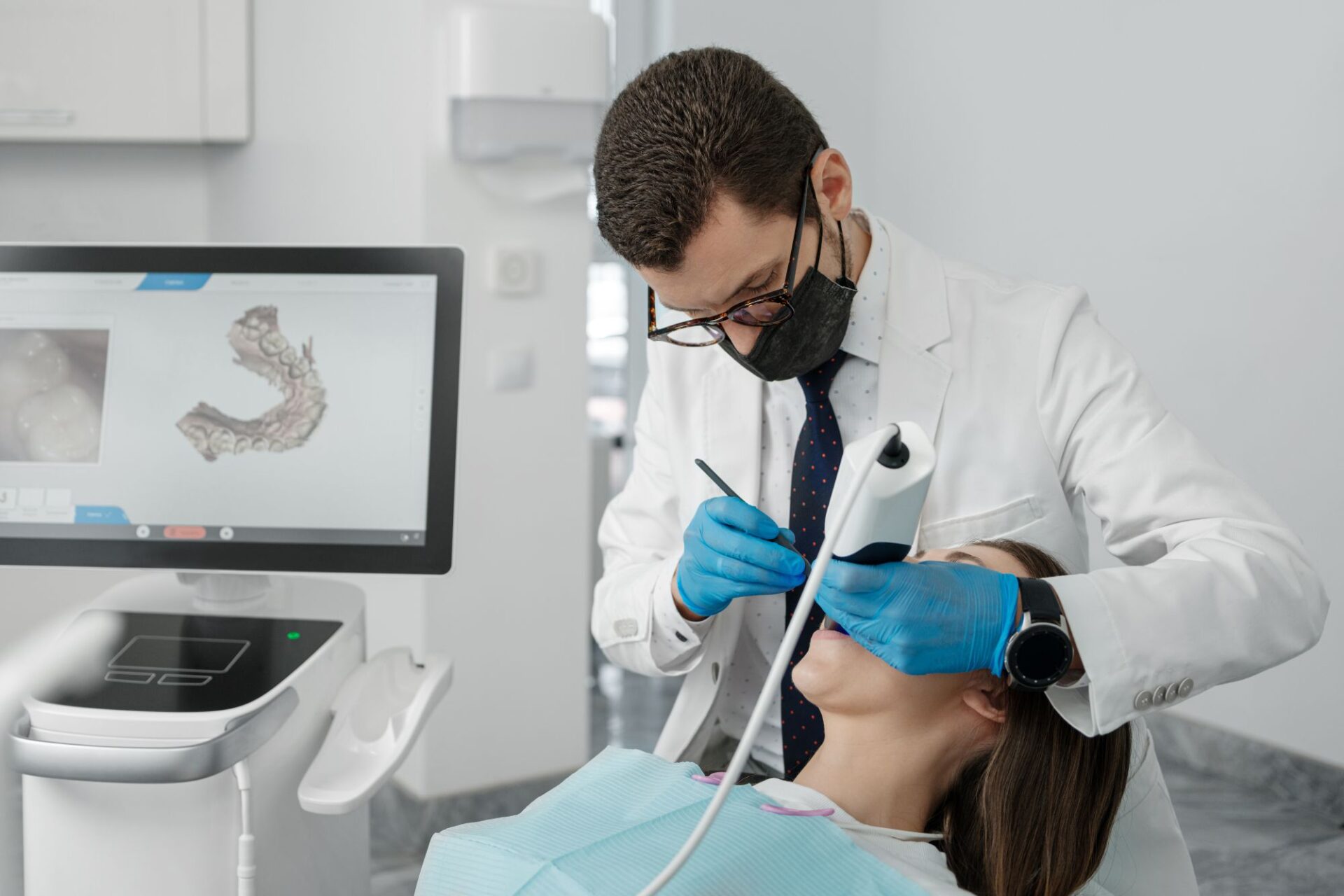 A dentist wearing a white coat, gloves, and a face mask is using an intraoral scanner to examine a female patient's teeth. A 3D rendering of the patient's dental structure is visible on the computer monitor, which is connected to the scanning device.