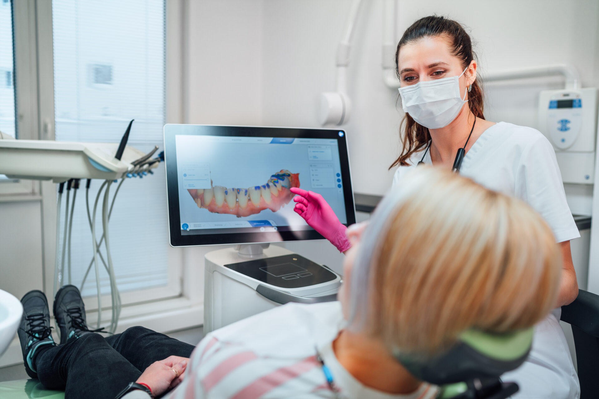 Dental professional wearing a mask and pink gloves showing a patient a 3D digital scan of teeth on a computer screen in a modern dental clinic, illustrating advanced imaging technology used for dental lab network collaboration.